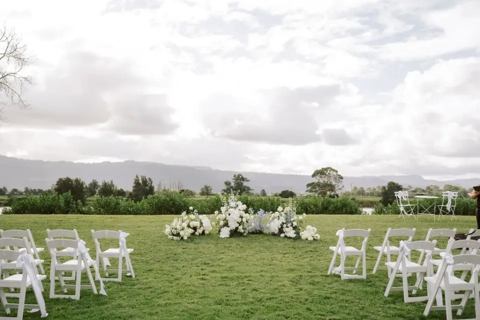 An empty ceremony lawn set with white chairs and a low white floral arrangement