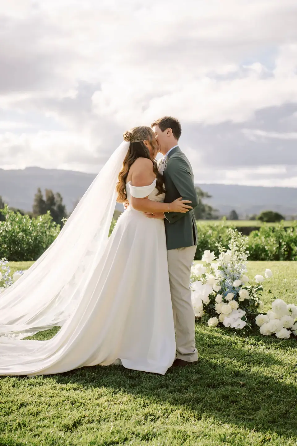 A couple's first kiss at an outdoor ceremony, the bride's veil trailing across the grass