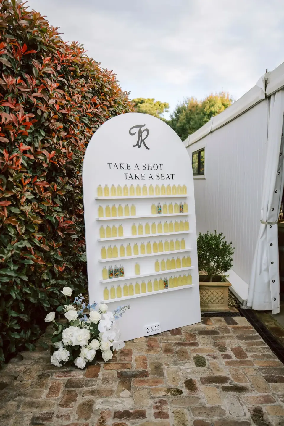 An arched wedding sign reading Take a shot, take a seat, holding a guest's drink for every seat